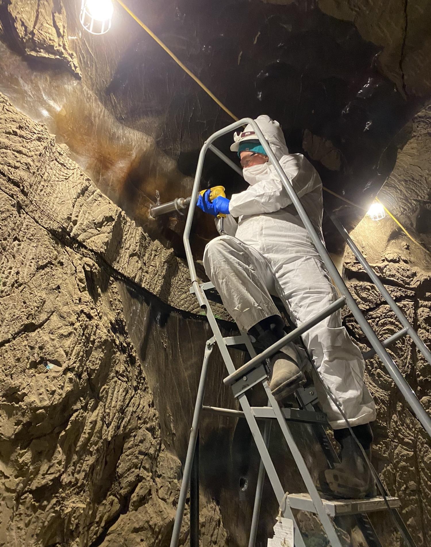 Man in full body suit stands on a ladder and drills into adjacent wall