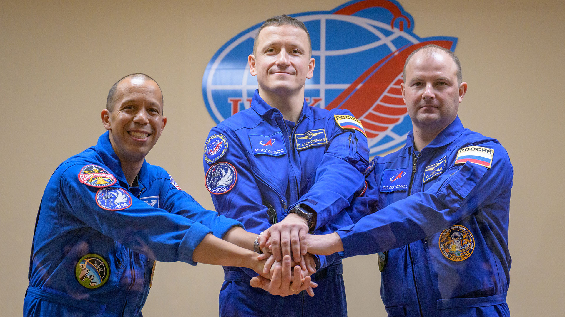 three men dressed in blue coveralls with colorful patches pose together with their hands stacked in front of them
