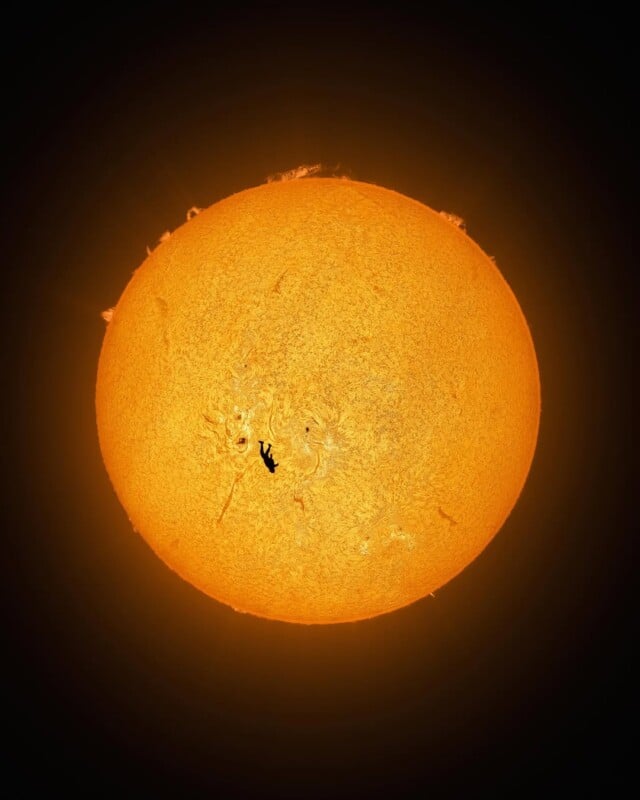 A bright, detailed image of the Sun with vivid orange and yellow surface textures. A small, dark silhouette of the International Space Station is visible crossing in front of the Sun.