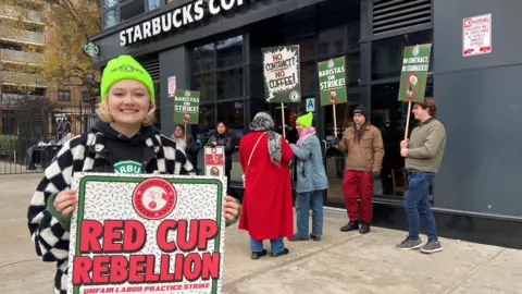 Danielle Kaye A woman wearing a neon beanie holds a sign reading "Red Cup Rebellion", standing in front of a Starbucks Coffee store alongside other people holding signs.