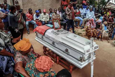 AFP via Getty Images Mourners gather around the coffin with the Chadema party flag laid on top to pay their final respects. The white coffin with silver handles is raised on a platform. 