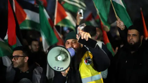 Reuters Pro-Palestinian protesters gather outside the stadium before the match. A man with a hi-vis jacket has a loudspeaker. The group are carrying flags.