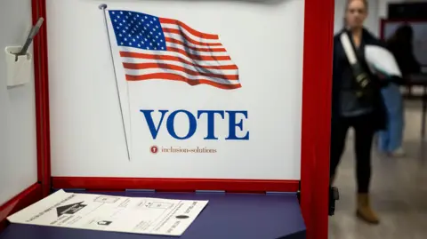 EPA/Shutterstock A blue table is blocked off by a white and red VOTE privacy screen. There is a ballot lying on the table and there appear to be people in the background in rack focus.