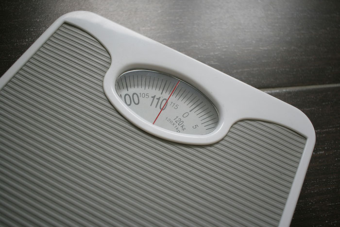 White analog bathroom scale showing a weight near 110 pounds on a dark wooden floor background.