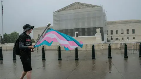 Getty Images Man in black T-shirt, black shorts and black hat carries flag with blue, pink and white stripes on rainy day with Supreme Court building, wrapped in light scaffolding in the background