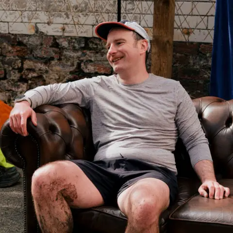 Xander Xander, a 37 year old man is sat on a on a sofa in shorts and a running top. He's laughing and he's got a muddy leg