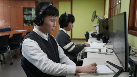 BBC/Hosu Lee Students wearing over-ear headphone sit at desks in front of computers screens and use screen readers and braille