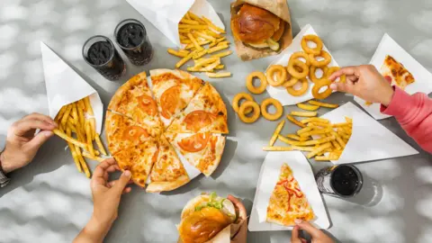 Getty Images Looking down on a table full of plates of pizza, burgers in buns, onion rings, french fries and cola drinks, which are all examples of ultra-processed foods. Hands are reaching out and picking up the different foods.