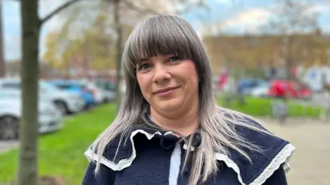 BBC A woman with grey hair is standing outside. She is wearing a navy top. The background is out of focus, but trees, grass and a variety of parked cars can be seen.