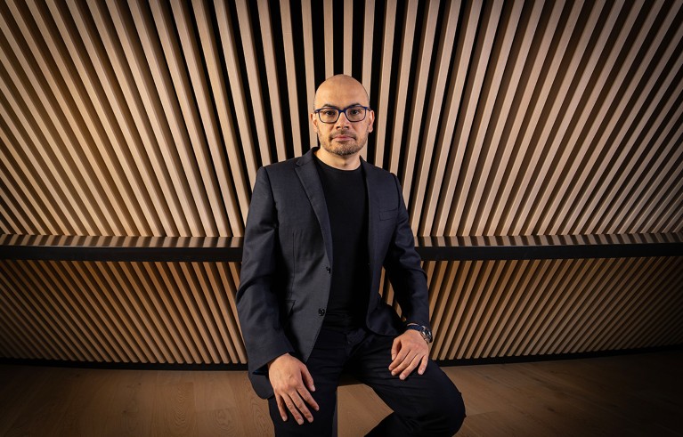 Portrait of Demis Hassabis in a black suit, seated, with a corrugated wooden backdrop.