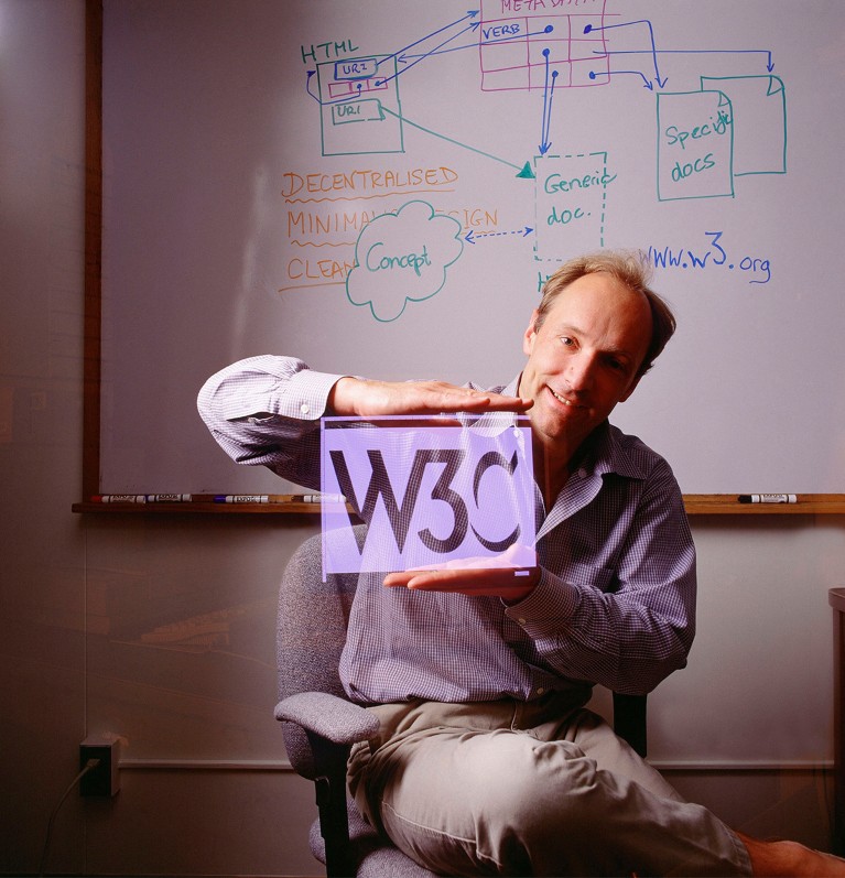 Tim Berners-Lee, British computer scientist and principal developer of the World Wide Web sitting in his office with a whiteboard behind him while holding a W3C lilac sign.