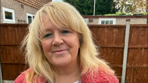 Heather Matuozzo has shoulder-length blonde hair and is wearing a pink top and is smiling at the camera.  She is standing in a garden with a brown fence behind her.
