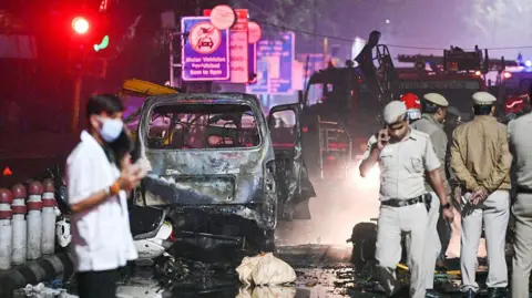 Getty Images Police officers and officials walk around the nighttime scene of a deadly car blast in India's capital city, Delhi. Behind them is the charred wreck of a burnt out car and a fire engine. The road is wet from firefighting efforts and covered in debris.