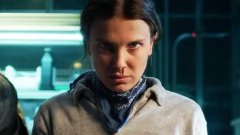 Netflix Stranger Things character Eleven, a teenager girl, is stood glaring directly at the camera. She is wearing a grey jumper with a navy, patterned neckerchief visible around her neck. A drip of blood is running from her nose towards her lip. Her head is lowered with her dark hair tied behind her head. Behind her, metallic shelves are visible, lit by neon lights and stacked with what appears to be bottles.