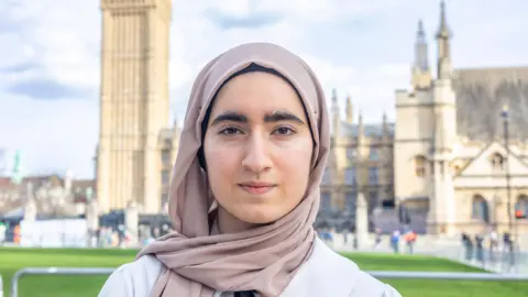 Kimi Gill Isra, a young woman wearing a head scarf, wears a neutral expression, in Parliament square, with the House of Parliament in the background