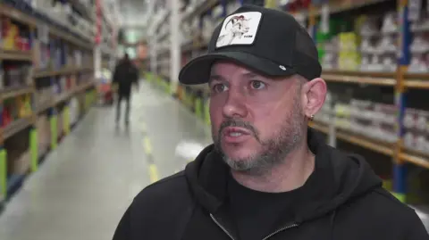 Comedian White Yardie outside a grocery shop, wearing a cap, black T-Shirt and black hoodie