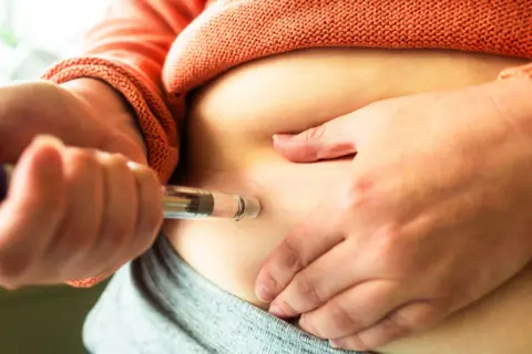 Getty Images Close up of a woman's hands holding a weight loss injection pen. She is injecting the dose into her abdomen.