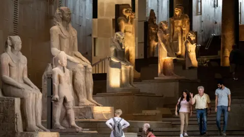 Getty Images A grand staircase displays statues of ancient kings and queens of Egypt, as visitors admire them.