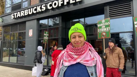 Danielle Kaye A woman wearing a neon beanie stands in front of a Starbucks coffee shop in Brooklyn, New York, alongside people holding signs reading "Baristas on Strike".