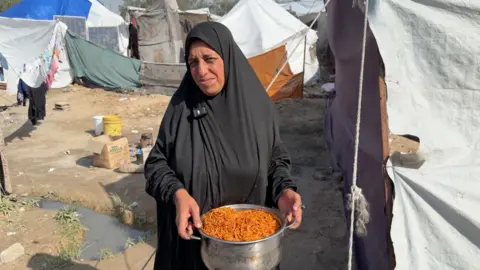 Aida Salha holds a pan full of pasta from an Anera community kitchen in central Gaza. Tents can be seen around her and she is wearing a black dress and headscarf