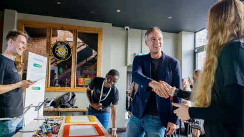 Google A group of people are gathered inside a modern, well-lit venue with large windows and exposed brick walls. On the right of the frame the UK government's busienss secretary, Peter Kyle MP, shakes hand with a blonde-haired woman. Next to them is a table covered with colourful LEGO pieces and two large baseplates. To the left, a person wearing a black Google-branded T-shirt stands near a tall display board titled “AI Quick Build,” which lists steps and information. Another person in a similar T-shirt is leaning forward, appearing to engage with the table setup.