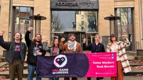 A group of six people are shown with their fists raised in the air, holding a banner for IWGB Game Workers Union that reads: "Game Workers Beat Bosses". They are standing in front of the Rockstar North office in Edinburgh.