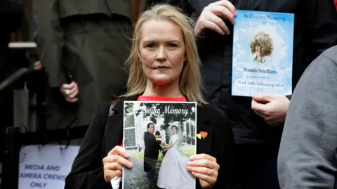 PA Media Catriona Myles holding a picture of a man and woman, she has blonde hair. She is wearing a black coat and red top.