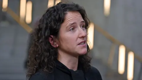 Getty Images Mairi Gougeon, who has brown curly hair, stands side on to the camera. She is wearing a dark top and is visible from the shoulders up. A concrete wall and steps, with wooden bannisters and lights are out of focus in the background. 