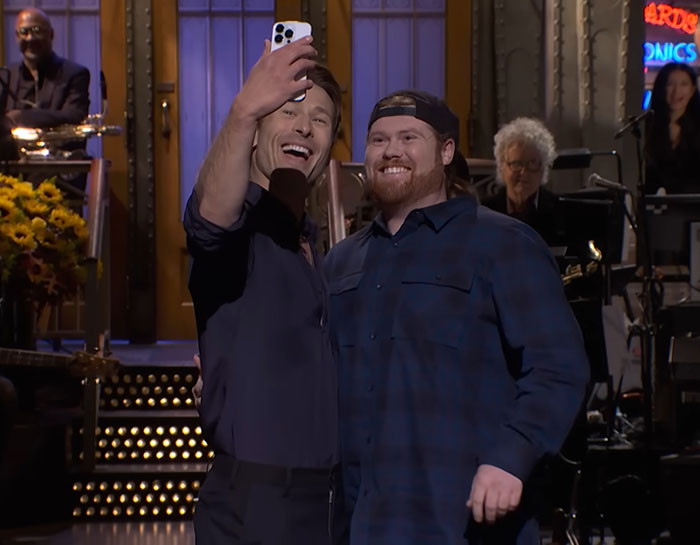 Glenn Powell smiling and taking a selfie with a UPS driver on stage during an emotional SNL moment. Glenn Powell smiling and taking a selfie with a UPS driver on stage during an emotional SNL moment.