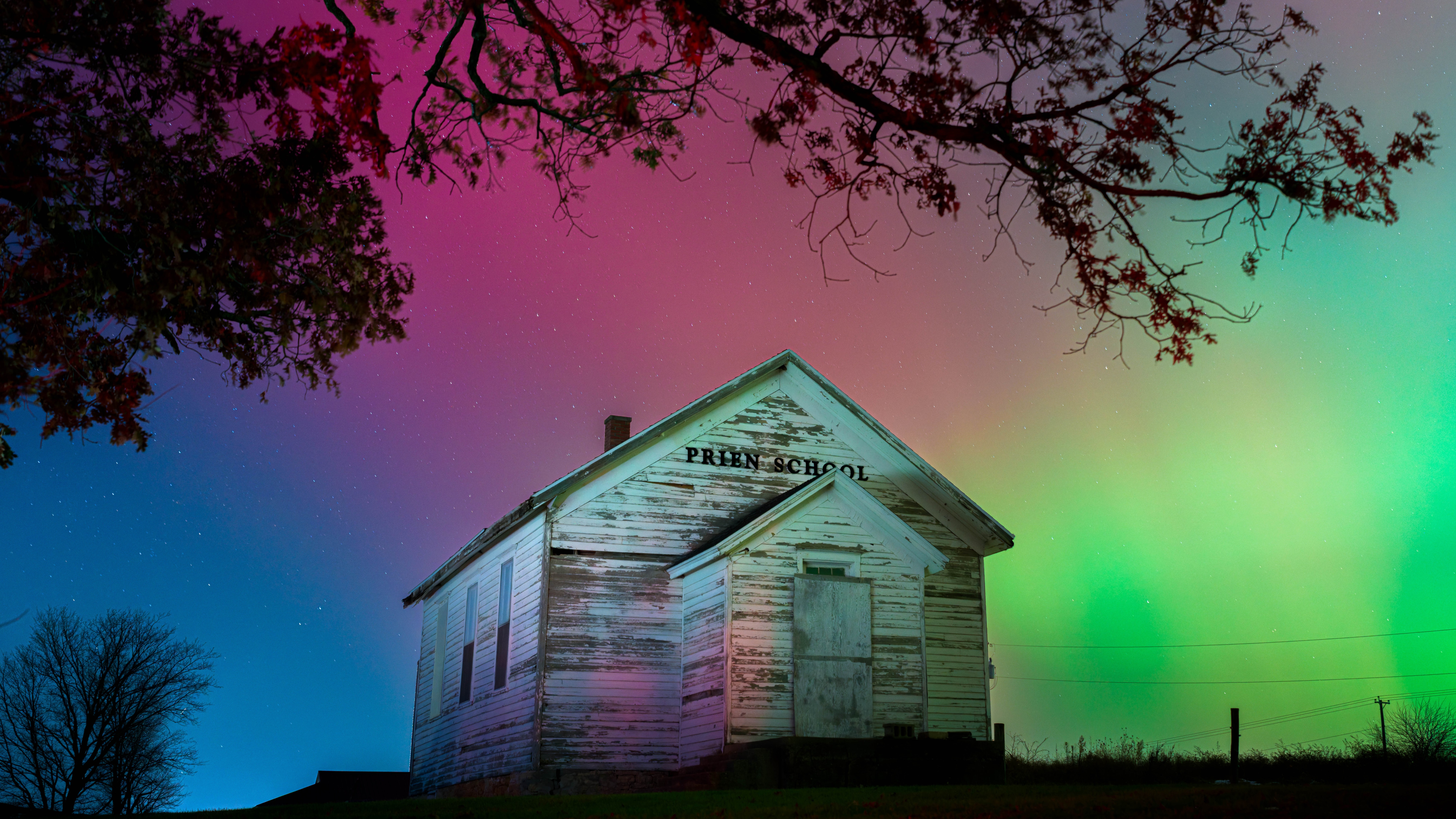 green and magenta aurora fill the sky with an old wooden rural building in the center of the image and a tree silhouetted in the foreground.