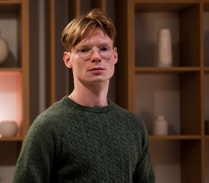 Young man with glasses and green sweater standing indoors in a room, reflecting mysteries about the human body science hasn't explained.