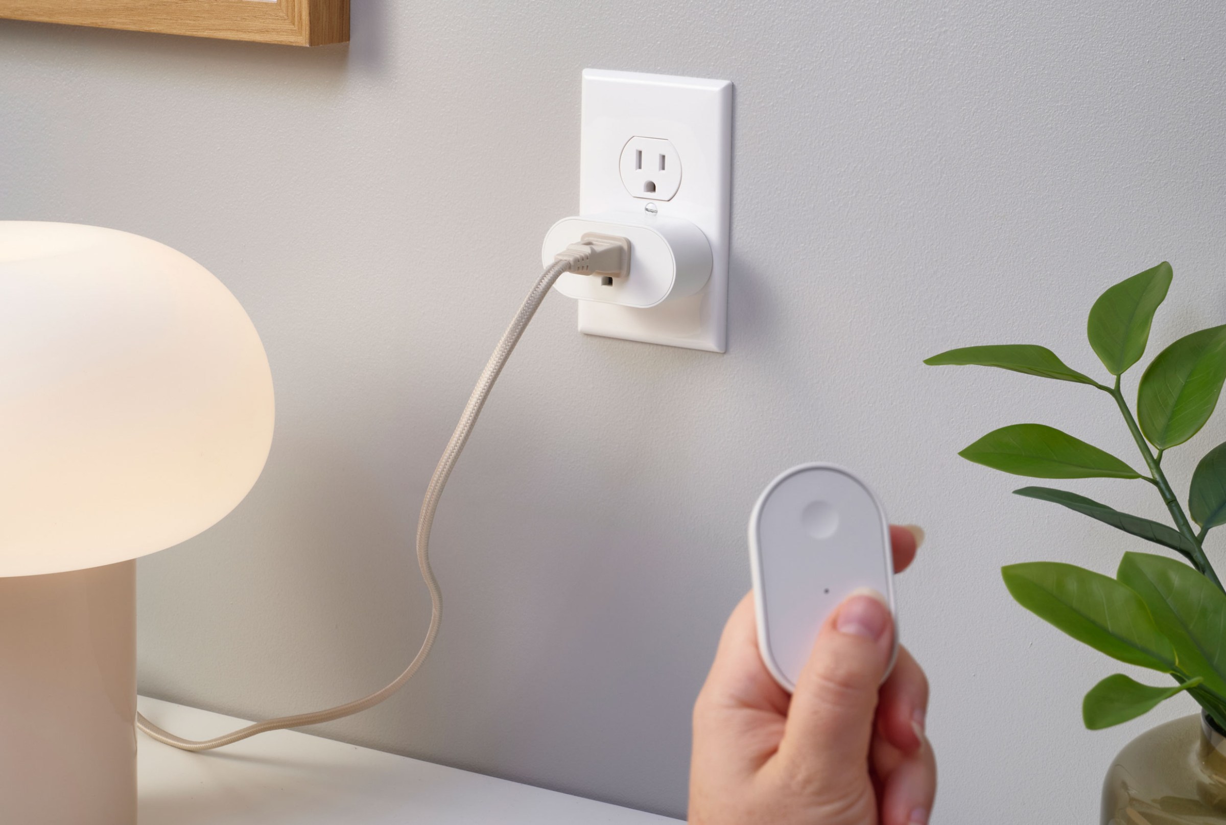 A hand holding a remote near a lamp plugged into a smart outlet adapter.