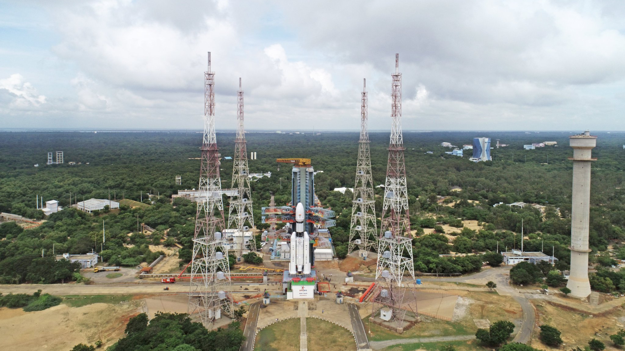 long-distance view of a white rocket on a launch pad, surrounded by green trees and shrubs