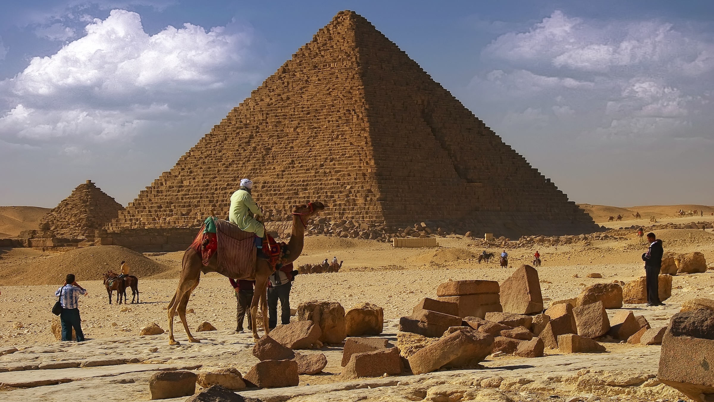 A few people and a camel are before a large Egyptian pyramid on a blue sky day.
