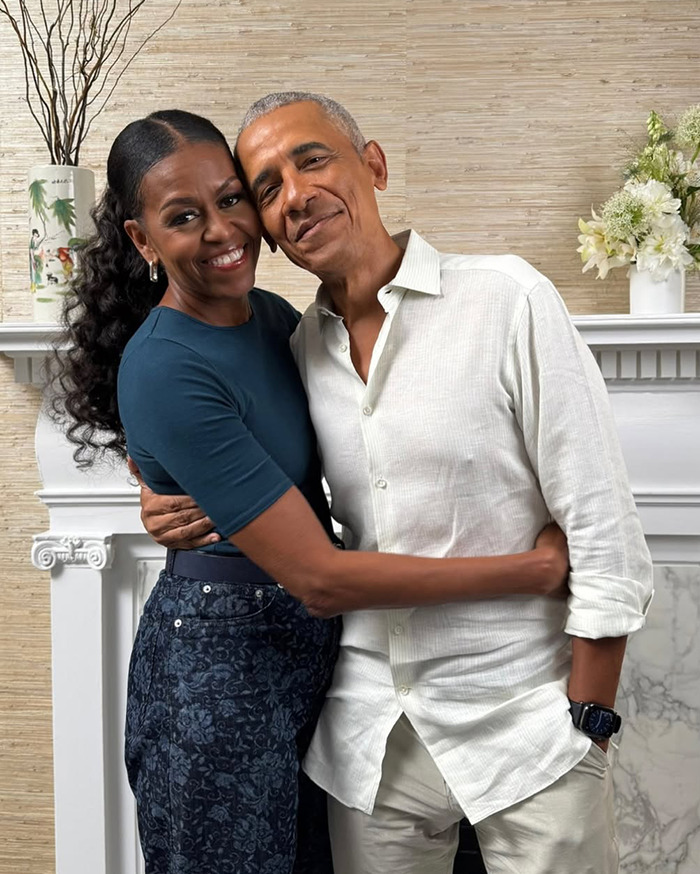 Michelle Obama smiling with ultra-long hair in a casual outfit, embracing Barack Obama indoors by a fireplace.