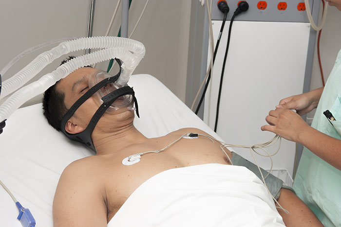 Man in hospital bed connected to breathing machine and medical monitors, representing coma survivors' stories and recovery.