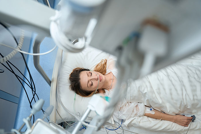 Young woman coma survivor lying in hospital bed connected to medical devices and monitoring equipment.