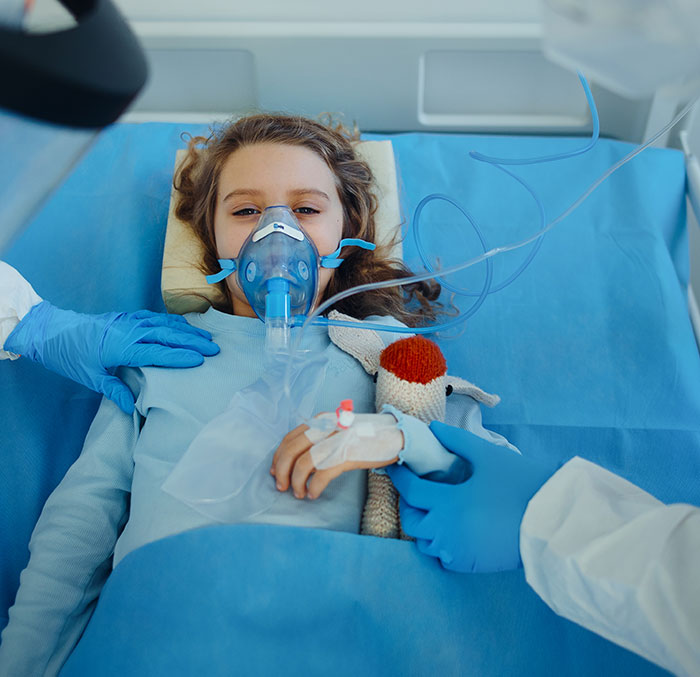 Young coma survivor girl in hospital bed with oxygen mask and IV, holding a stuffed animal, surrounded by medical staff.