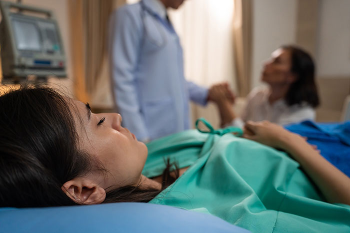 Young woman lying in hospital bed in coma while doctor and family hold hands in background, coma survivors story concept.