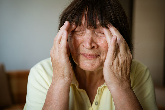 Elderly woman with closed eyes holding her head, reflecting the struggles faced by coma survivors sharing their stories.