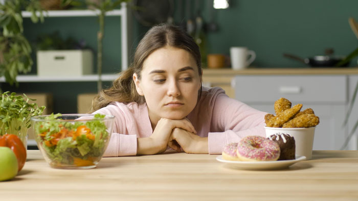 Young woman in kitchen choosing between fast food and healthy salad, reflecting on strict diet and eating disorder triggers. Young woman in kitchen choosing between fast food and healthy salad, reflecting on strict diet and eating disorder triggers.