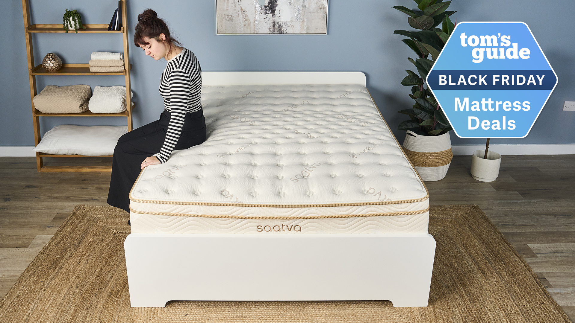 Mattress tester Lauren Jeffries sits on the side of the Saatva Classic mattress on a bed frame in a decorated bedroom, to test the edge support. In the top right corner, a Tom's Guide Black Friday mattress deals graphic