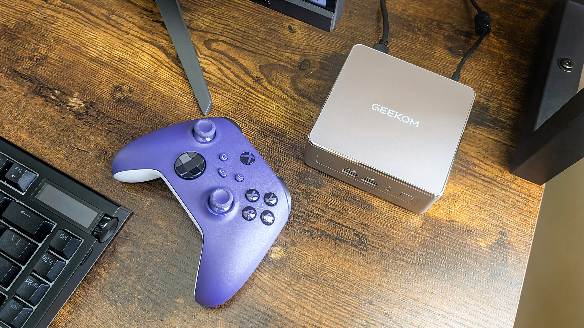 A purple Xbox Controller laying on a desk next to a Geekom mini PC, a keyboard, and a monitor.