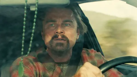 Warner Bros Leonardo DiCaprio in One Battle After Another, driving a car and looking concerned while glancing in the rear-view mirror