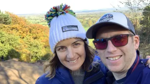 Andrew and Zoë Zoe and Andrew are photographed on a sunny day in the countryside. They are both wearing blue jackets. She has a white bobble hat; he is wearing a baseball cap.