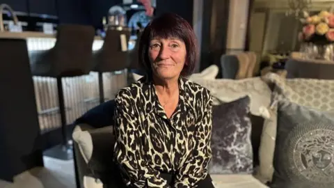 Debbie Matthews, a woman in a leopard print shirt with bobbed brown hair, sitting on a grey sofa.