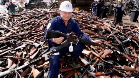 Getty Images A man, in a blue jumpsuit and wearing a white hardhat, crouching on top of a large pile of rifles while holding one 