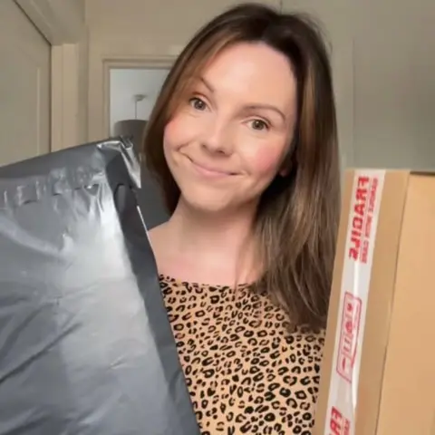 Kirsty Quinn Kirsty Quinn with shoulder length brown hair holding a parcel either side of her and smiling at the camera