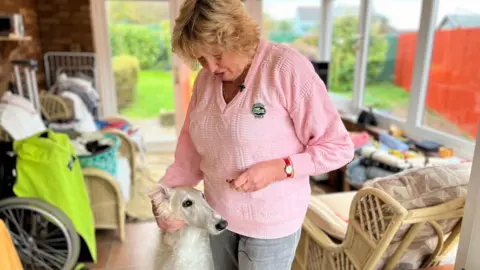 Mrs Gifford wearing a pink jumper with her arm around the head of her white Russian borzoi dog. They are standing in a conservatory surrounded by furniture.