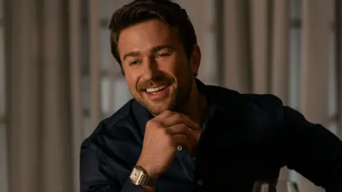 Lionsgate Actor Brandon Sklenar appearing in film The Housemaid. He is smiling whilst holding his chin with his hand. He is wearing a blue shirt.